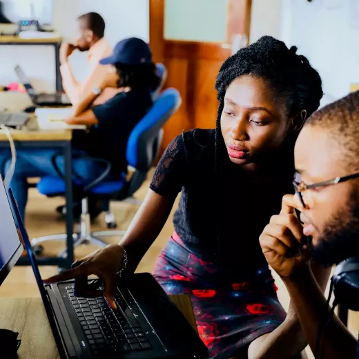 Two professionals, a woman and a man, closely examining a laptop screen in a busy office setting, showing focus and collaboration.