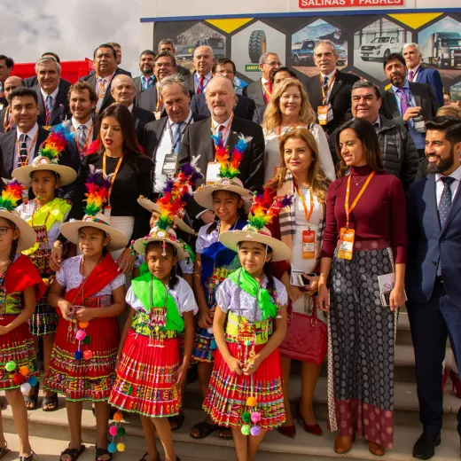 A group of adults in business attire and children in traditional colorful outfits with flower necklaces at an outdoor event, posing for a photo, expressing cheerful demeanors.