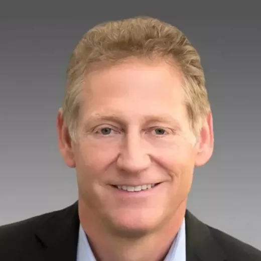 Headshot of Kent Masters, CEO of Albemarle a smiling Caucasian man with light blonde hair, wearing a dark suit and a white shirt, against a gray background.