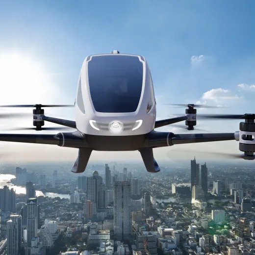 A futuristic drone or flying taxi with four rotors, featuring a sleek white cabin, hovers above a cityscape under a clear blue sky.