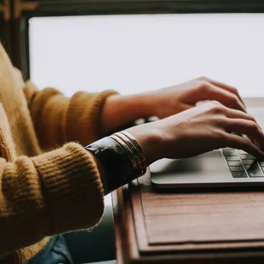 A person in a yellow sweater typing on a laptop with a strong, fire-resistant circuit board. The laptop is placed on a wooden desk, near a window with natural light streaming in.