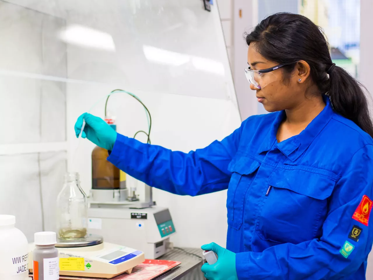 A scientist wearing a blue lab coat, safety glasses, and green gloves is working in a lab. She is handling a glass container at a lab station, surrounded by various lab equipment and bottles.
