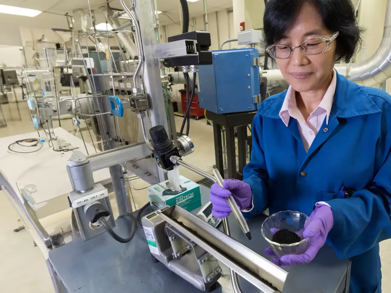 A female scientist in a lab coat conducts an experiment in an industrial setting, interacting with equipment while examining a sample in a mixing dish.