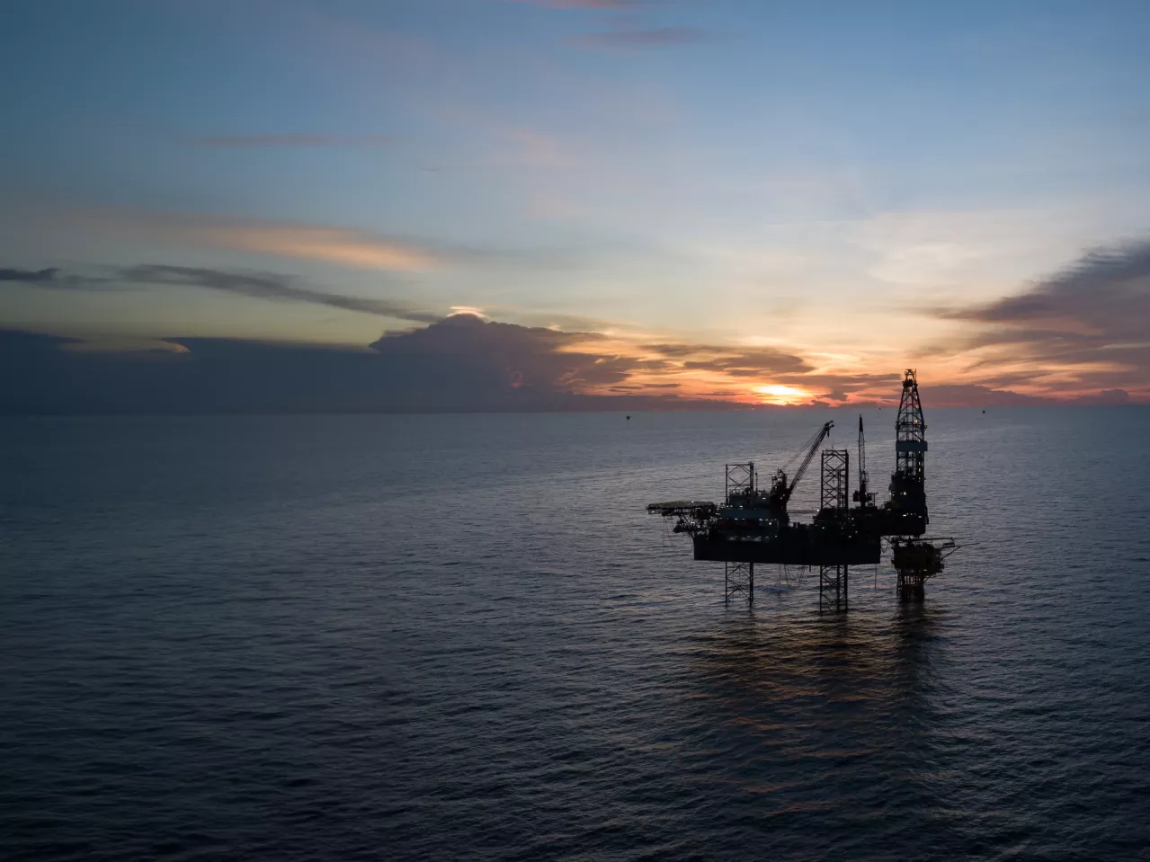An offshore oil rig silhouetted against a dramatic sunset sky over a calm ocean.